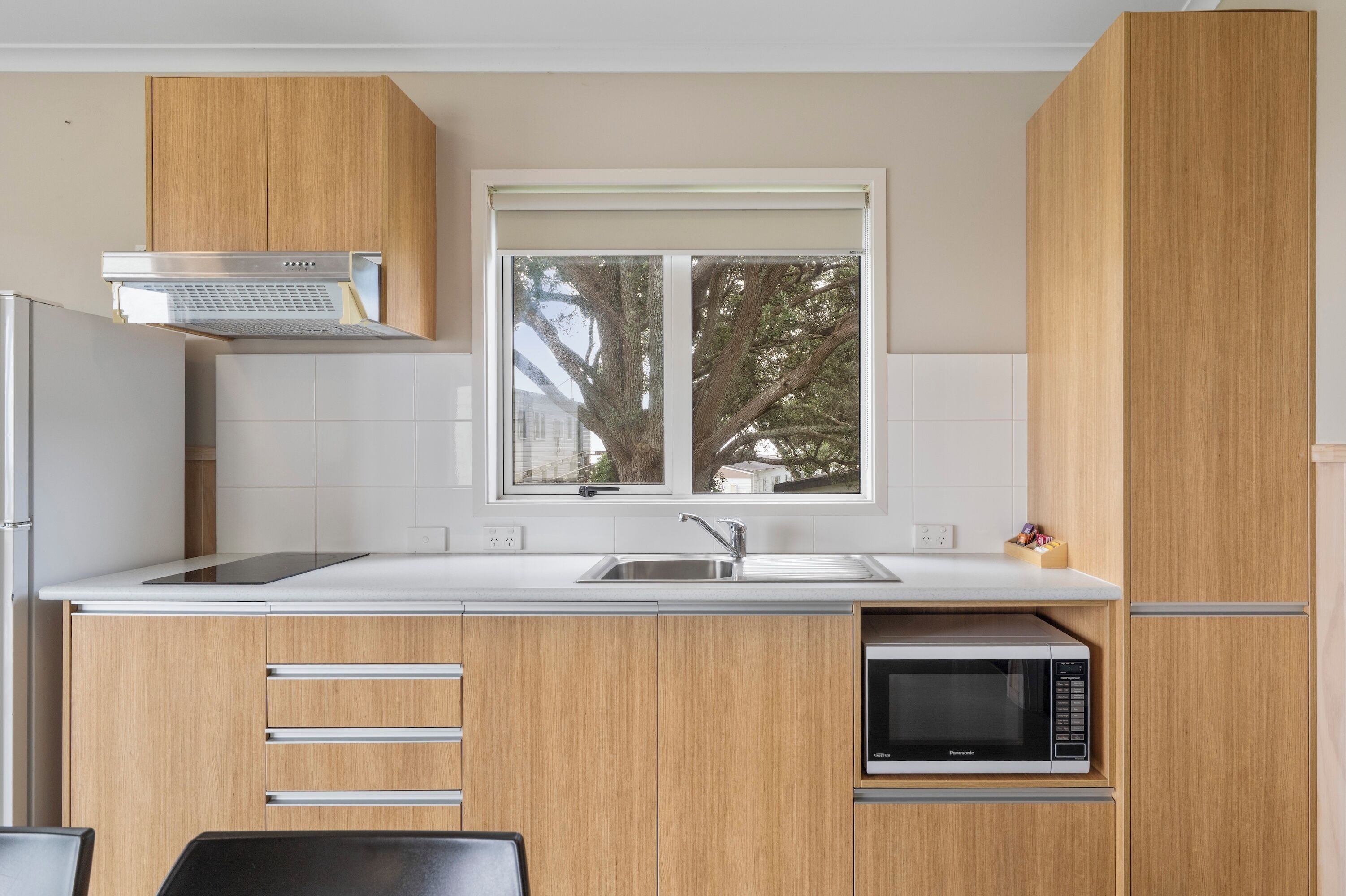 A compact kitchen with wooden cabinets, a stainless steel range hood, a built-in microwave, and a window with a view of trees.