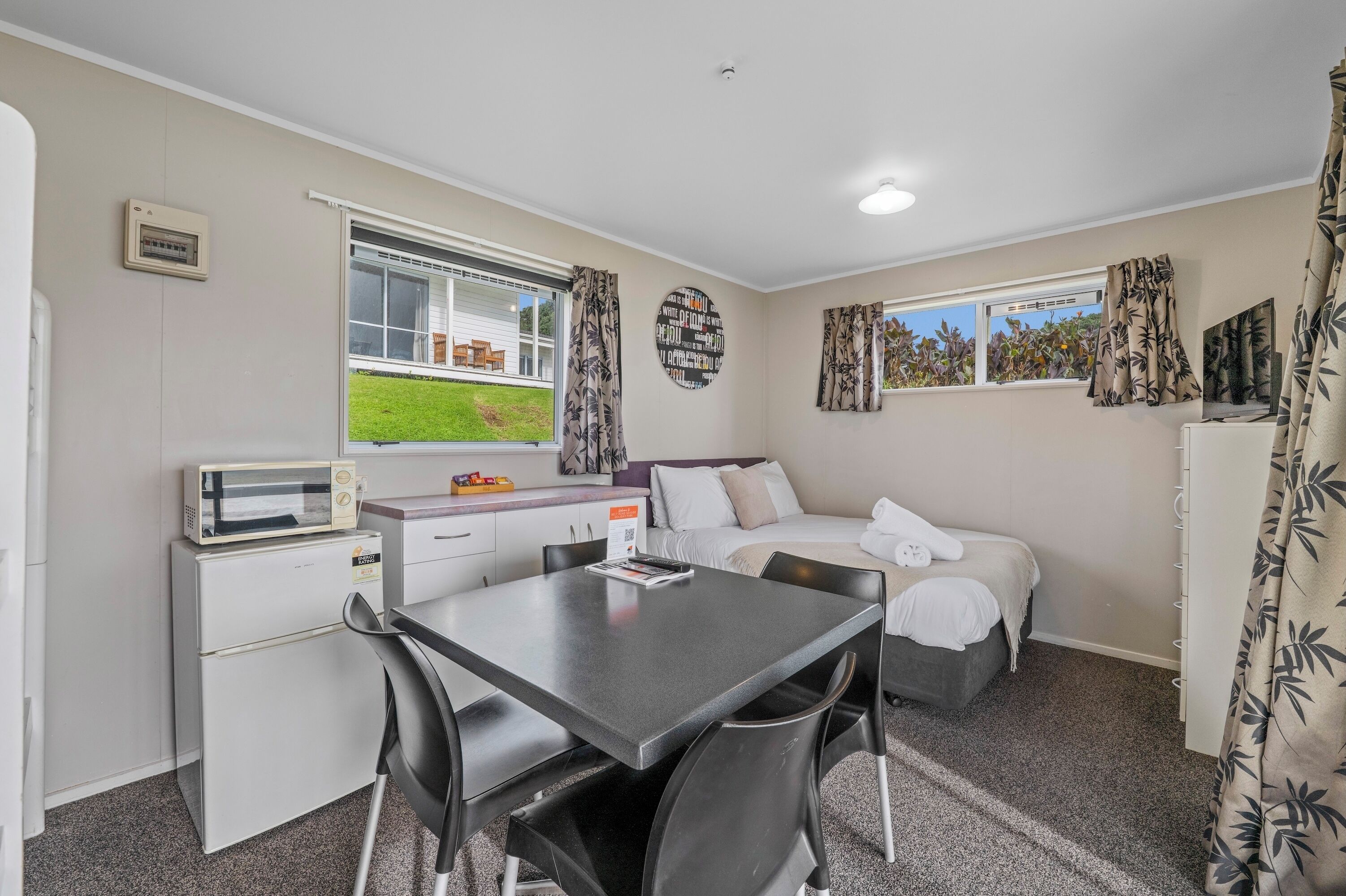 A compact room with a bed, a small kitchenette, a dining table, and a window with curtains.
