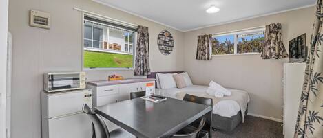 A compact room with a bed, a small kitchenette, a dining table, and a window with curtains.