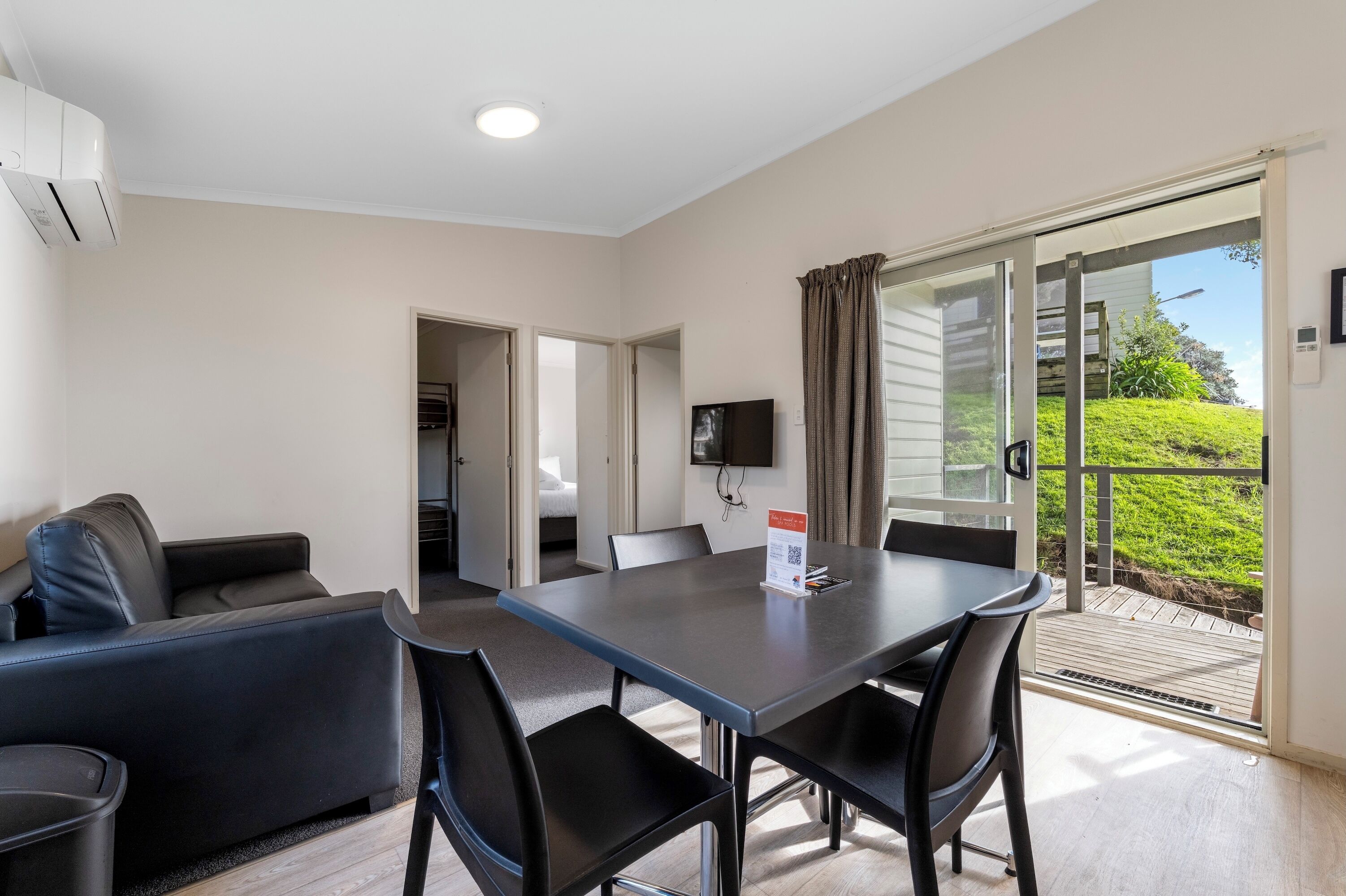 Habitación de hotel moderna con zona de comedor, sofá, televisor de pantalla plana y balcón con vistas a zonas verdes.
