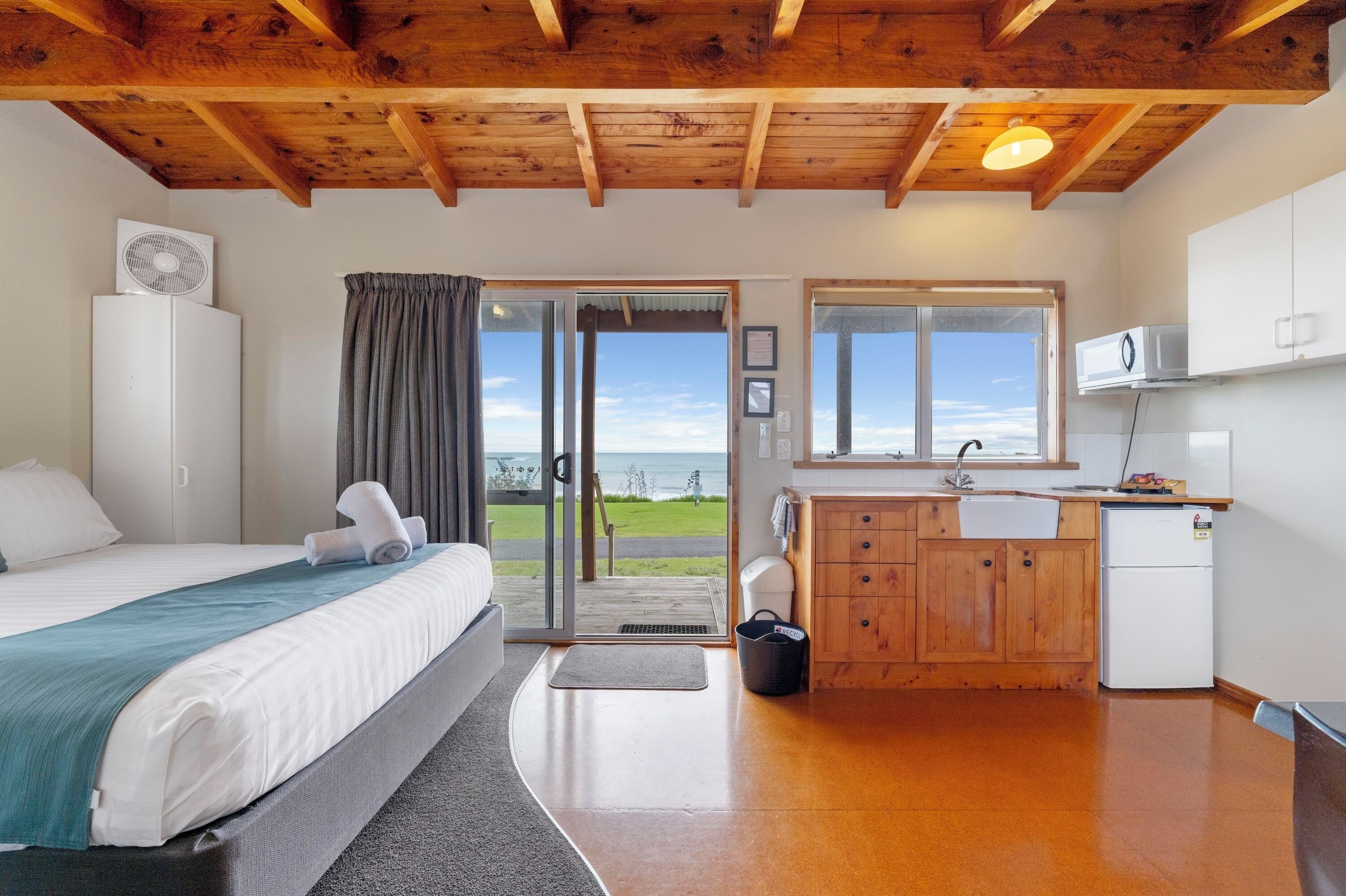 A bedroom with a bed, a fan, a view of the outdoors, a kitchen area with cabinets and a refrigerator, and a wooden ceiling.