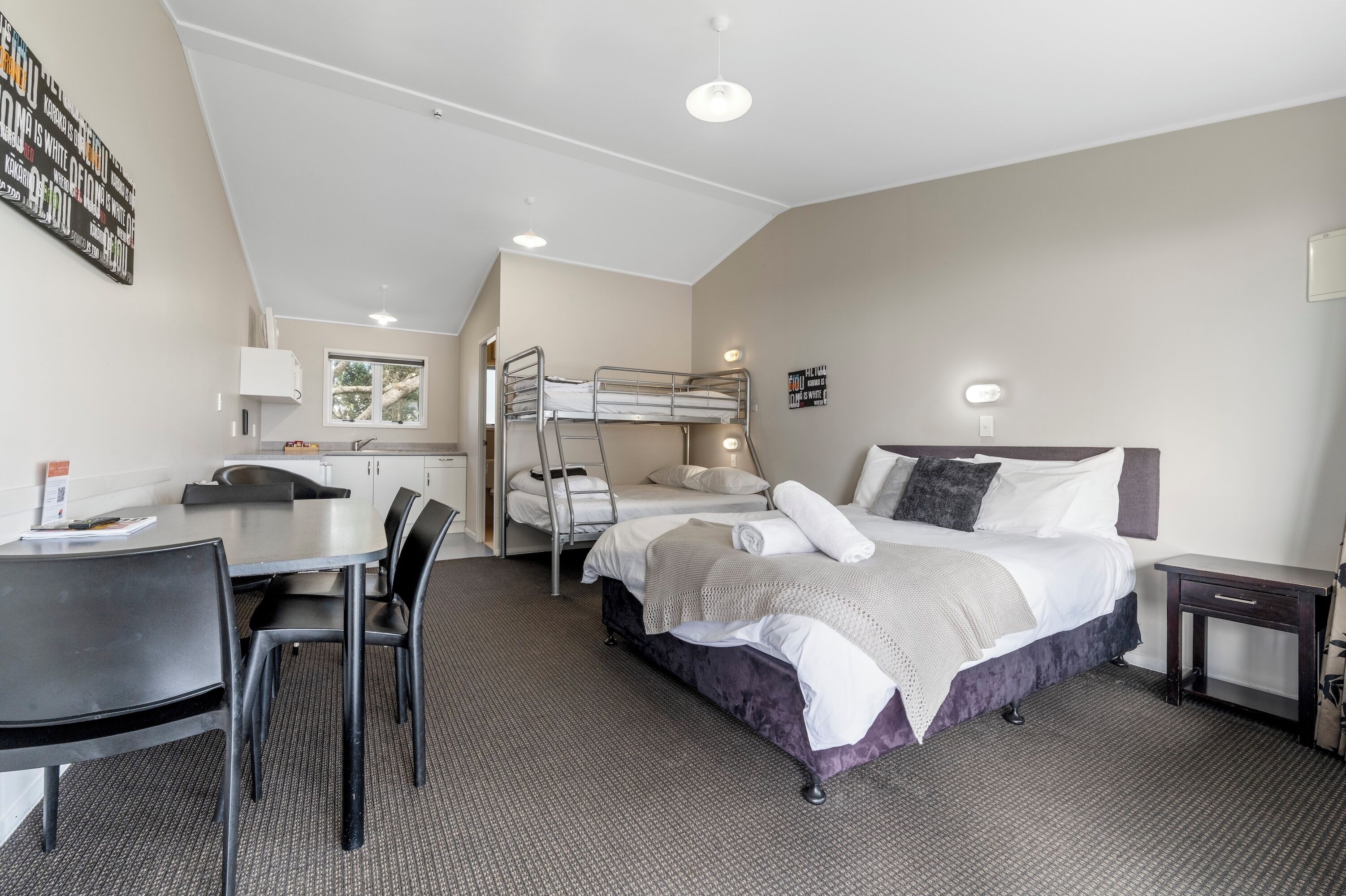 A hotel room with a bunk bed, a desk with chairs, a small table, and a bedside table.