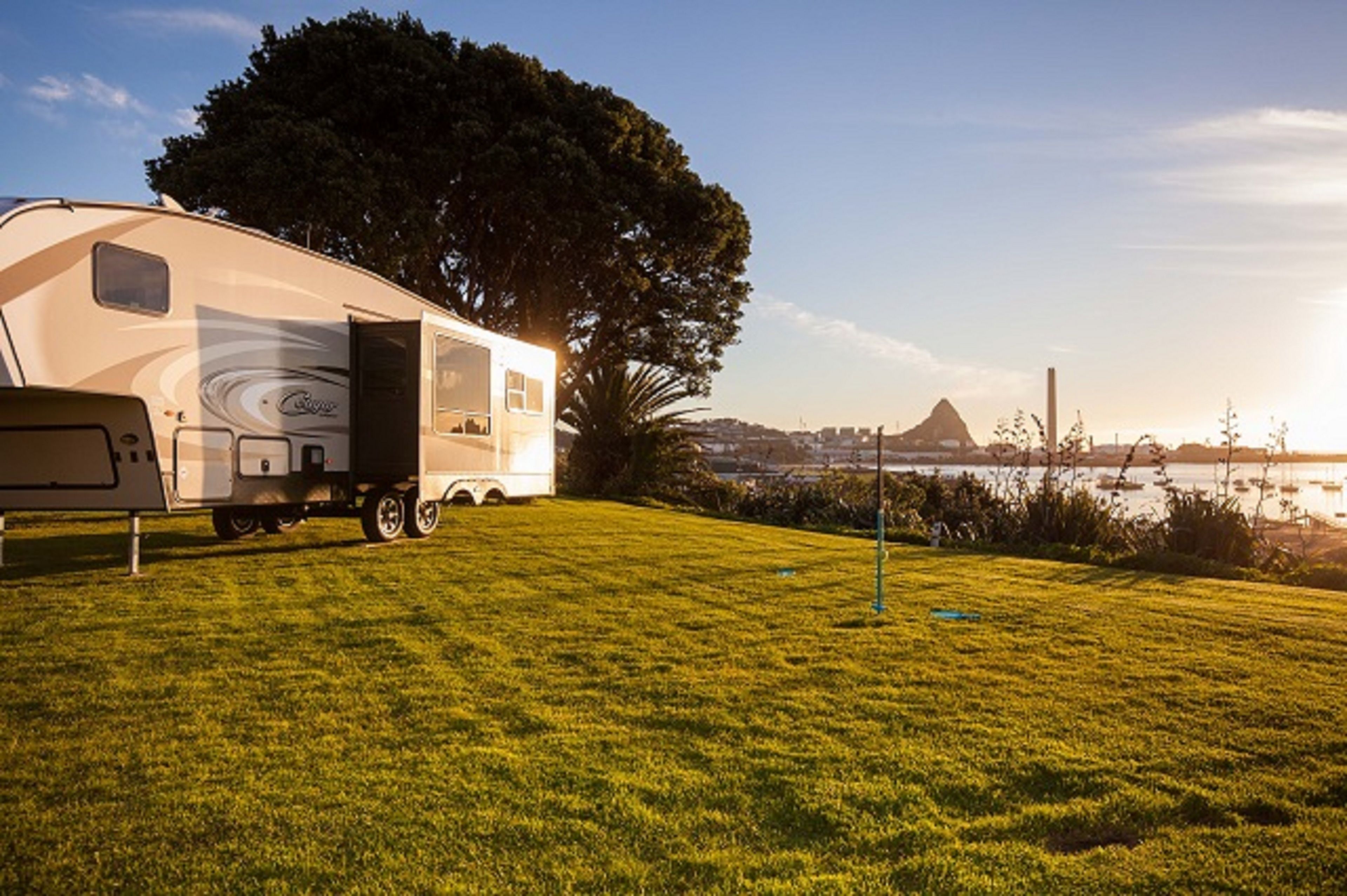 A recreational vehicle parked on a grassy area with a large tree and a view of a distant mountain and water body.