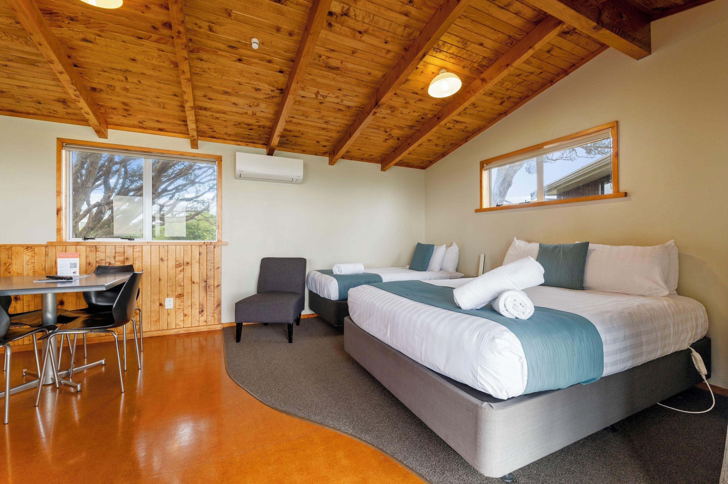 A room with a wooden ceiling, two beds with white and teal bedding, a small desk with a chair, and a window with a view of trees.