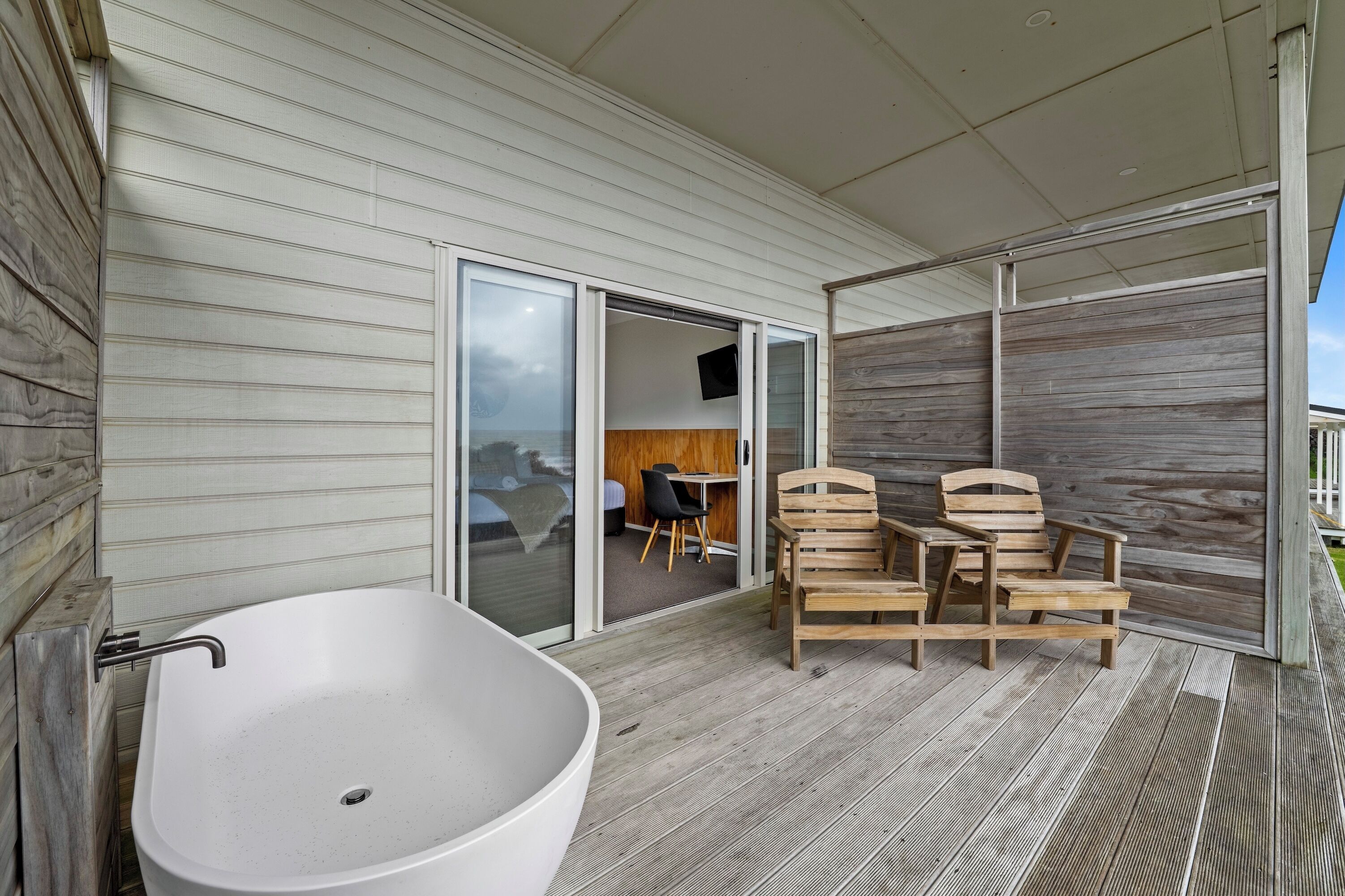 A modern bathroom with a freestanding bathtub, wooden deck, and a sliding glass door leading to a bedroom.