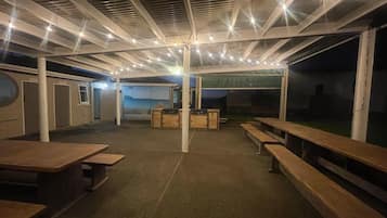 An outdoor covered area with wooden benches and tables, a grill station, and a view of the sea.
