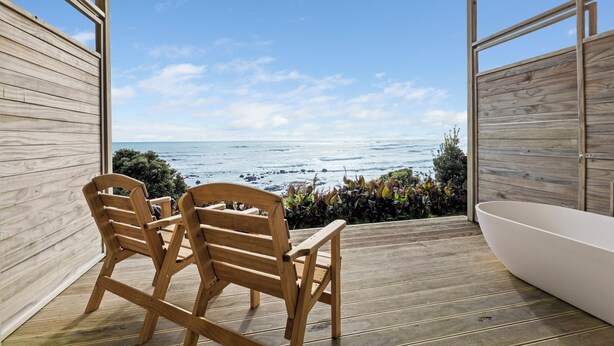 Une terrasse en bois avec vue sur l’océan, deux chaises en bois et une baignoire blanche.
