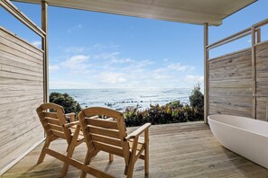 Terraza de madera con vista al océano, dos sillas de madera y una bañera blanca.