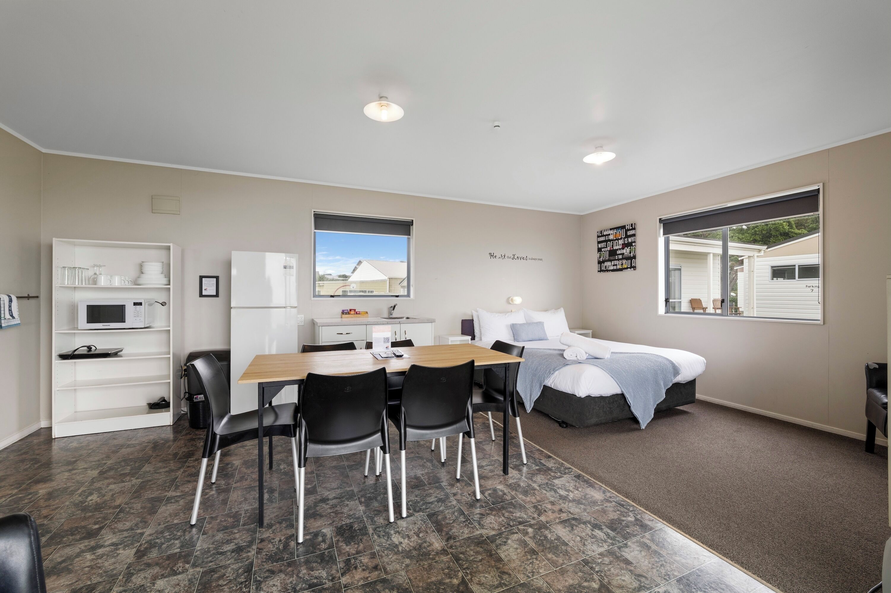 A compact hotel room with a kitchenette, a dining area, a bed, and a large window.