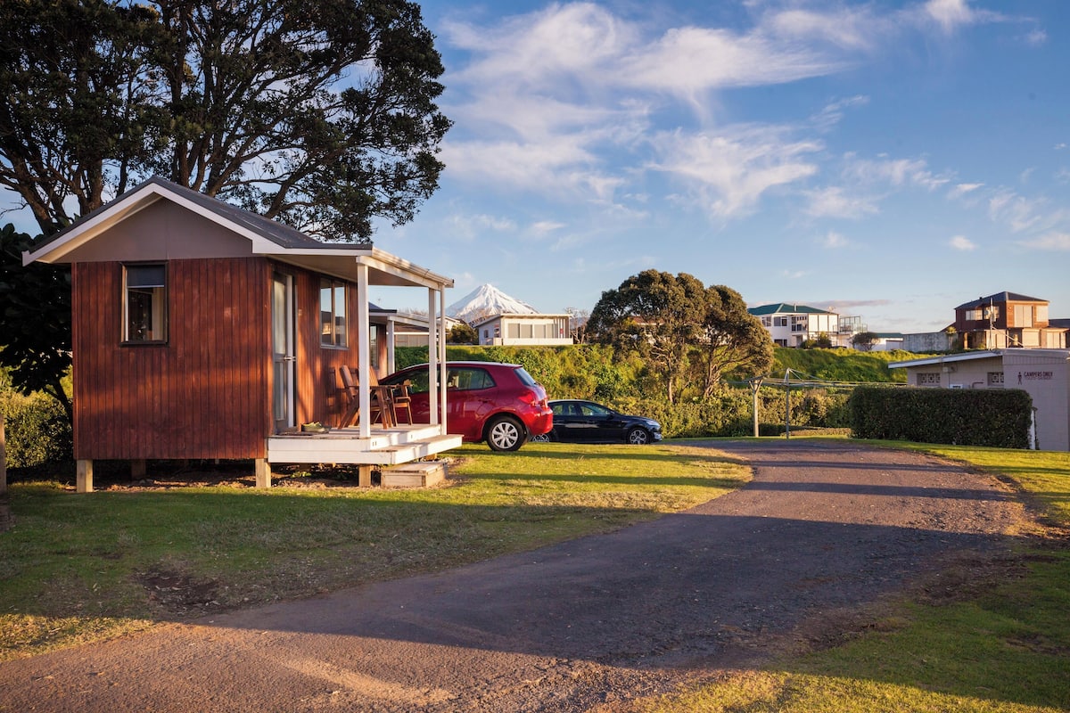 Eine kleine Holzhütte mit Veranda, davor ein rotes Auto und im Hintergrund ein Berg.