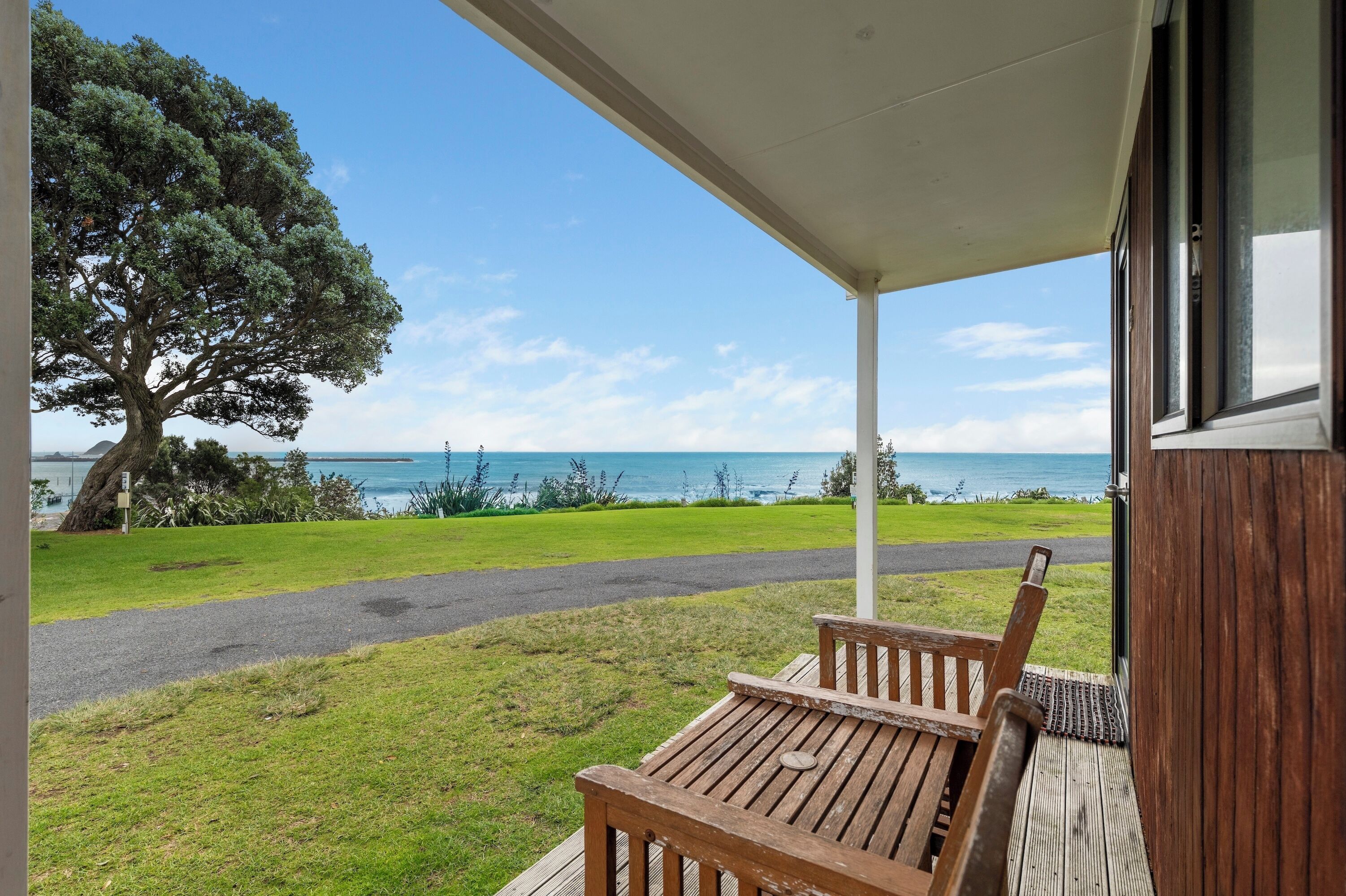Una terraza de madera con un banco que da a un área verde y vista al mar.