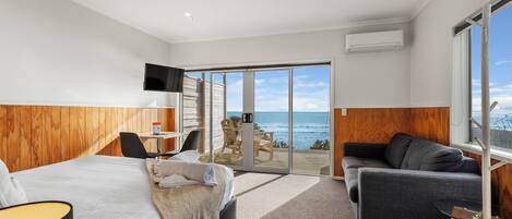 A modern hotel room with a large bed, a sofa, a desk, and a view of the ocean through glass doors.