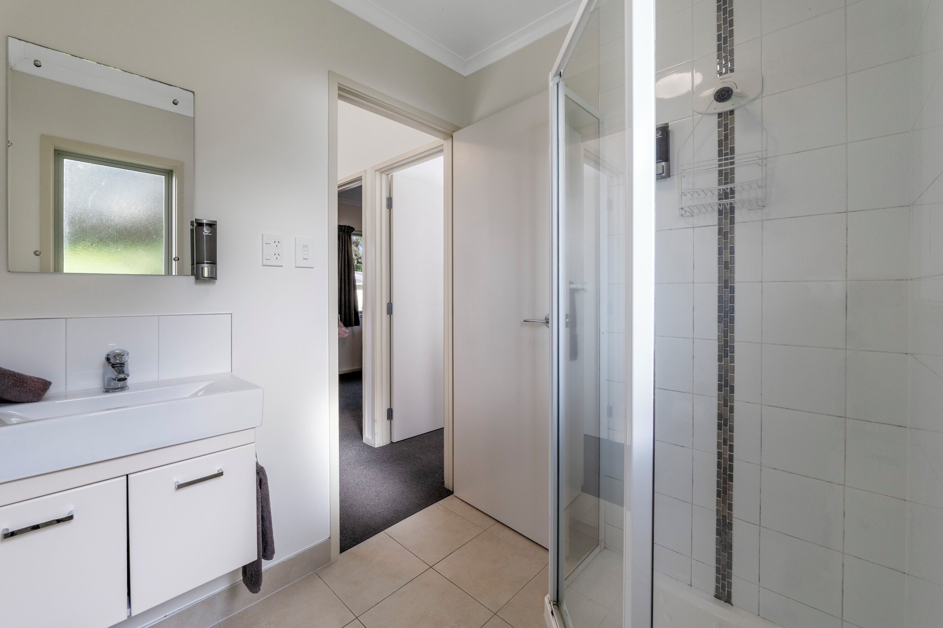 A modern bathroom with a glass shower enclosure, a white vanity with a mirror, and a door leading to another room.