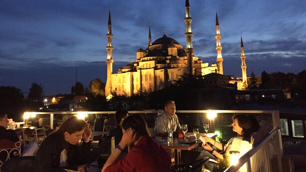 A group of people dining outdoors with a historic building featuring multiple minarets in the background. - Nobel Hostel Guesthouse (Istanbul)