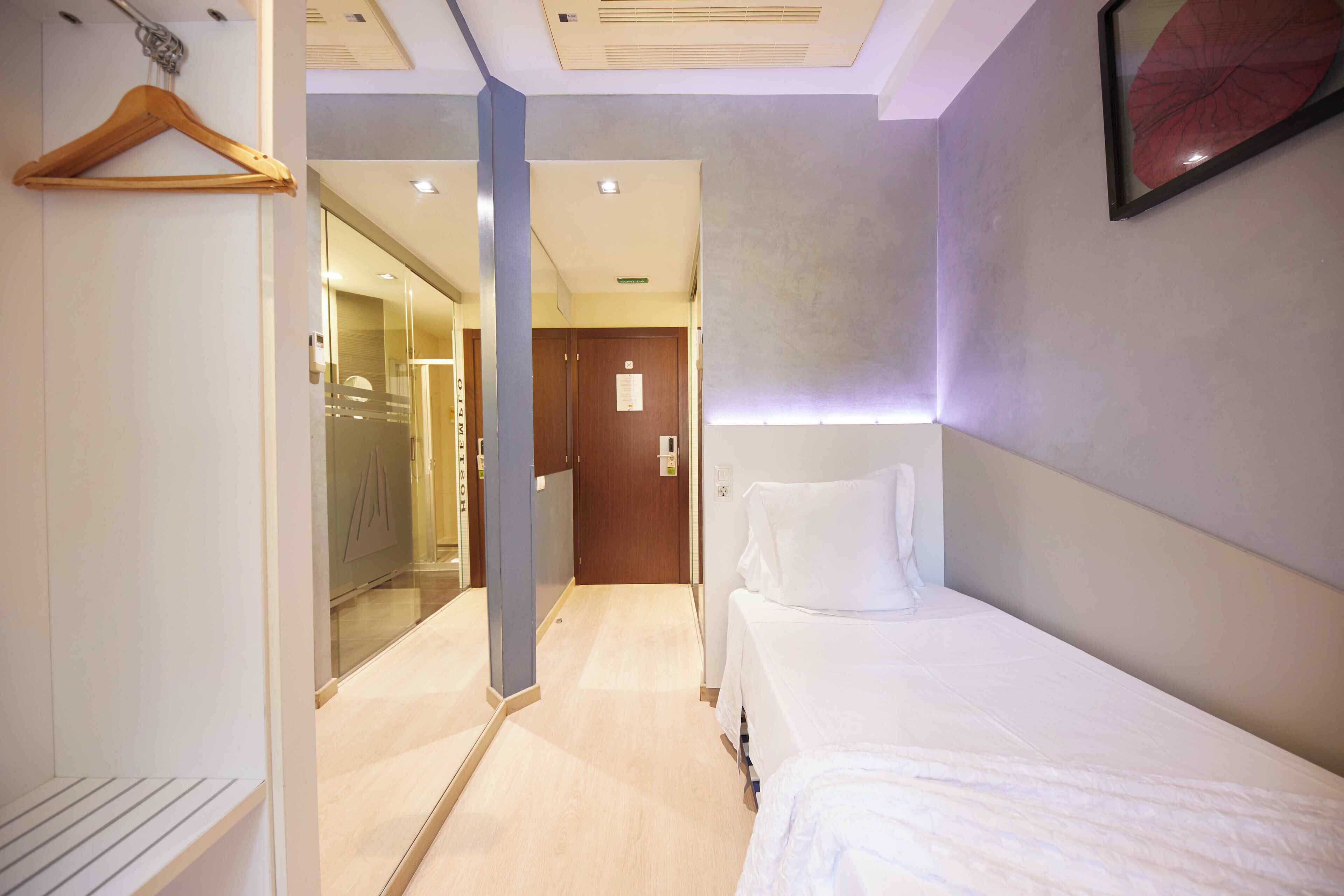 Una habitación de hotel con una cama, una percha de madera, un espejo, una puerta y un pasillo que conduce a un baño.