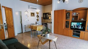 A living room with a sofa, a dining area with a table and chairs, a kitchen area with cabinets and a TV, and a wooden ceiling.