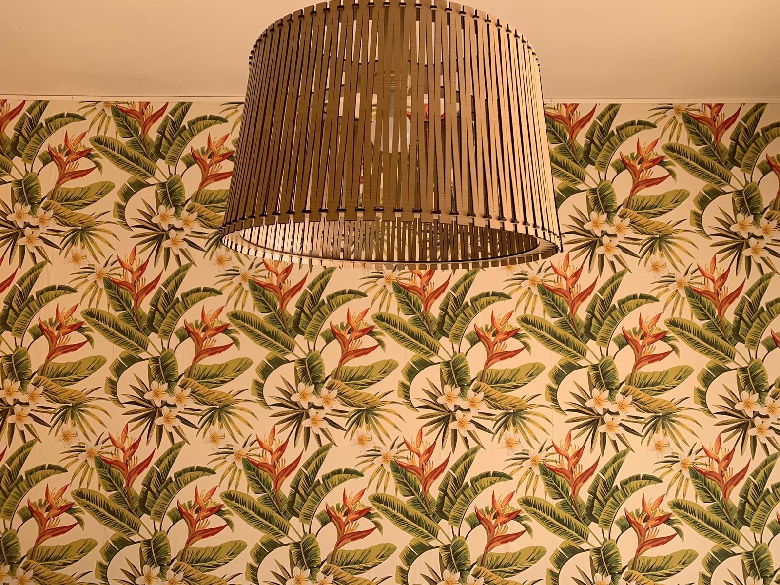 Una habitación con papel tapiz estampado con plantas y flores tropicales, una lámpara de techo con listones de madera y una pared lisa.