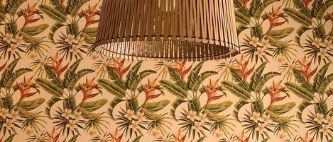 Une pièce dont le papier peint présente un motif avec des plantes et des fleurs tropicales, un lampadaire au plafond avec des lattes en bois, et un mur uni.