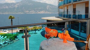 A balcony with a table, a drink, and a view of the sea and mountains. - Hotel Caribe by Double Hospitality (Brenzone sul Garda)