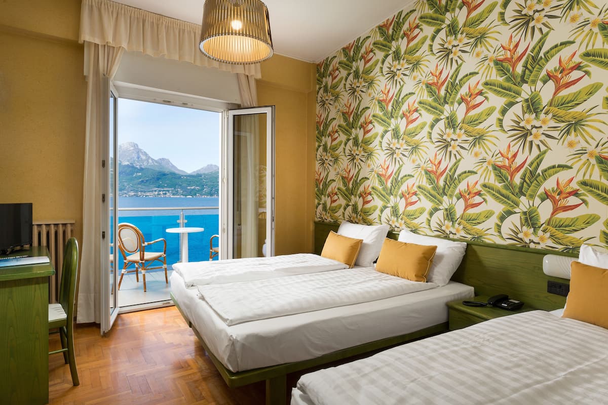 A hotel room with two beds, a desk, and a chair. There is a view of the sea and mountains through an open door.