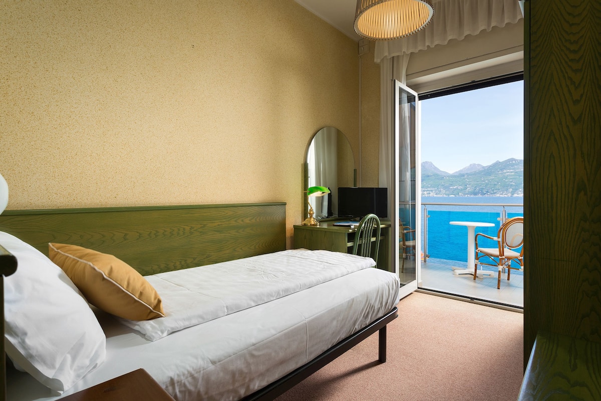 A hotel room with a bed, a desk, a chair, a mirror, and a view of the sea through a sliding door.