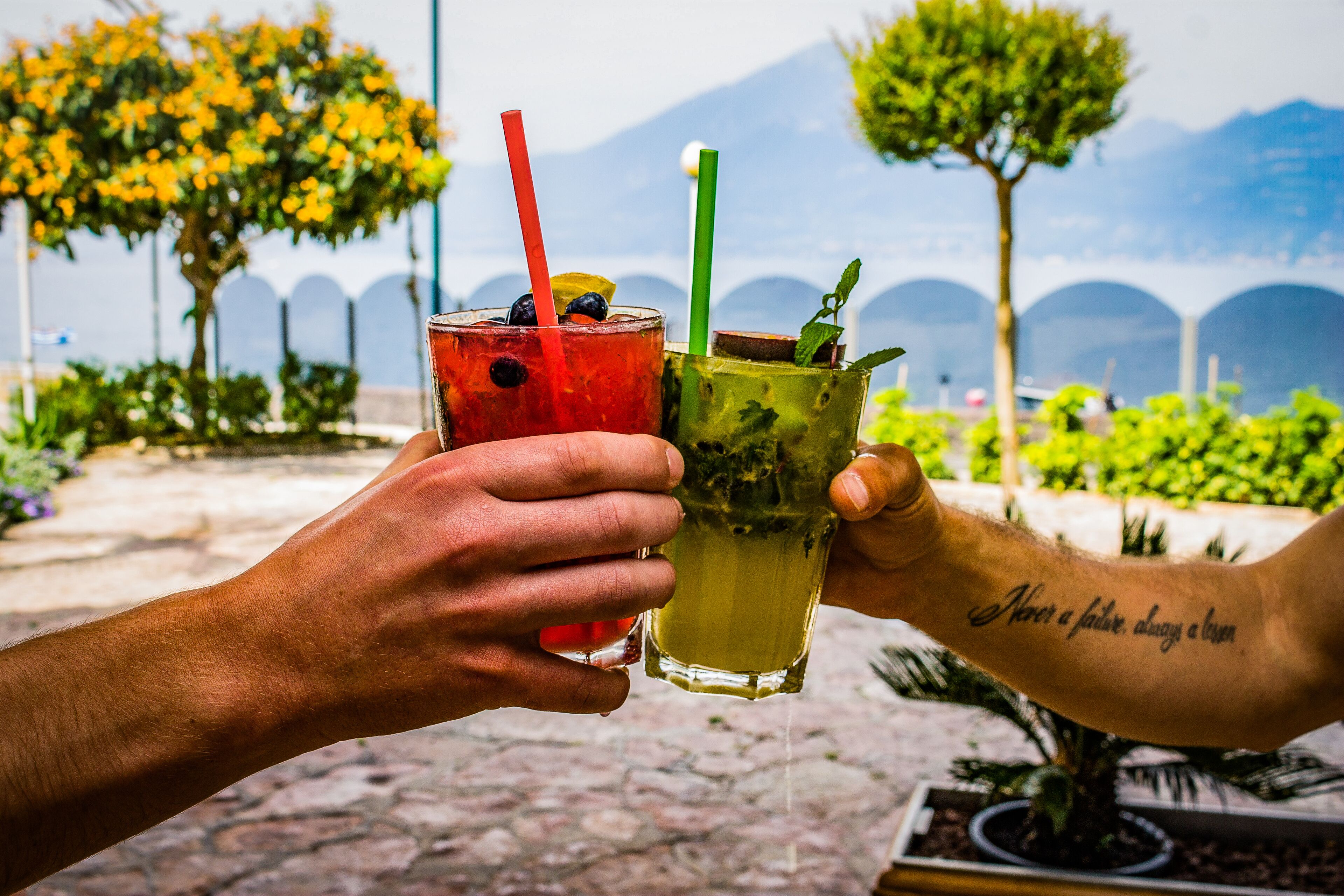 Two people holding glasses of drinks with straws, one with a red straw and one with a green straw, in an outdoor setting with trees and a distant view of mountains.
