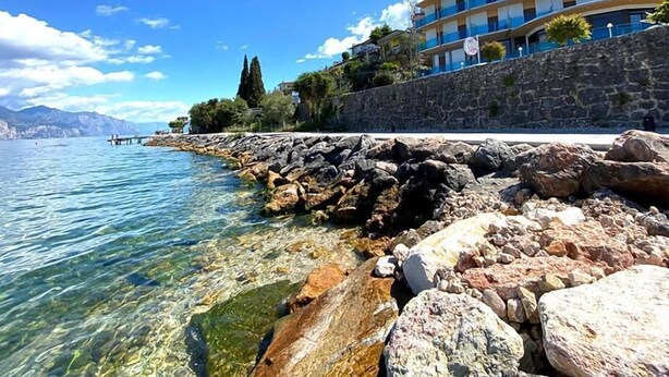 A coastal hotel with a stone wall and clear waters. - Hotel Caribe by Double Hospitality (Brenzone sul Garda)