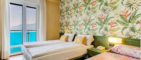 A hotel room with two beds, a view of the sea through a sliding door, and a tropical wallpaper.