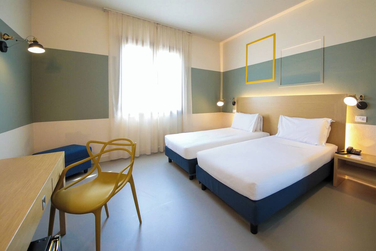 Ein Hotelzimmer mit zwei Betten, einem Schreibtisch, einem Stuhl und einem fenster mit Vorhängen.