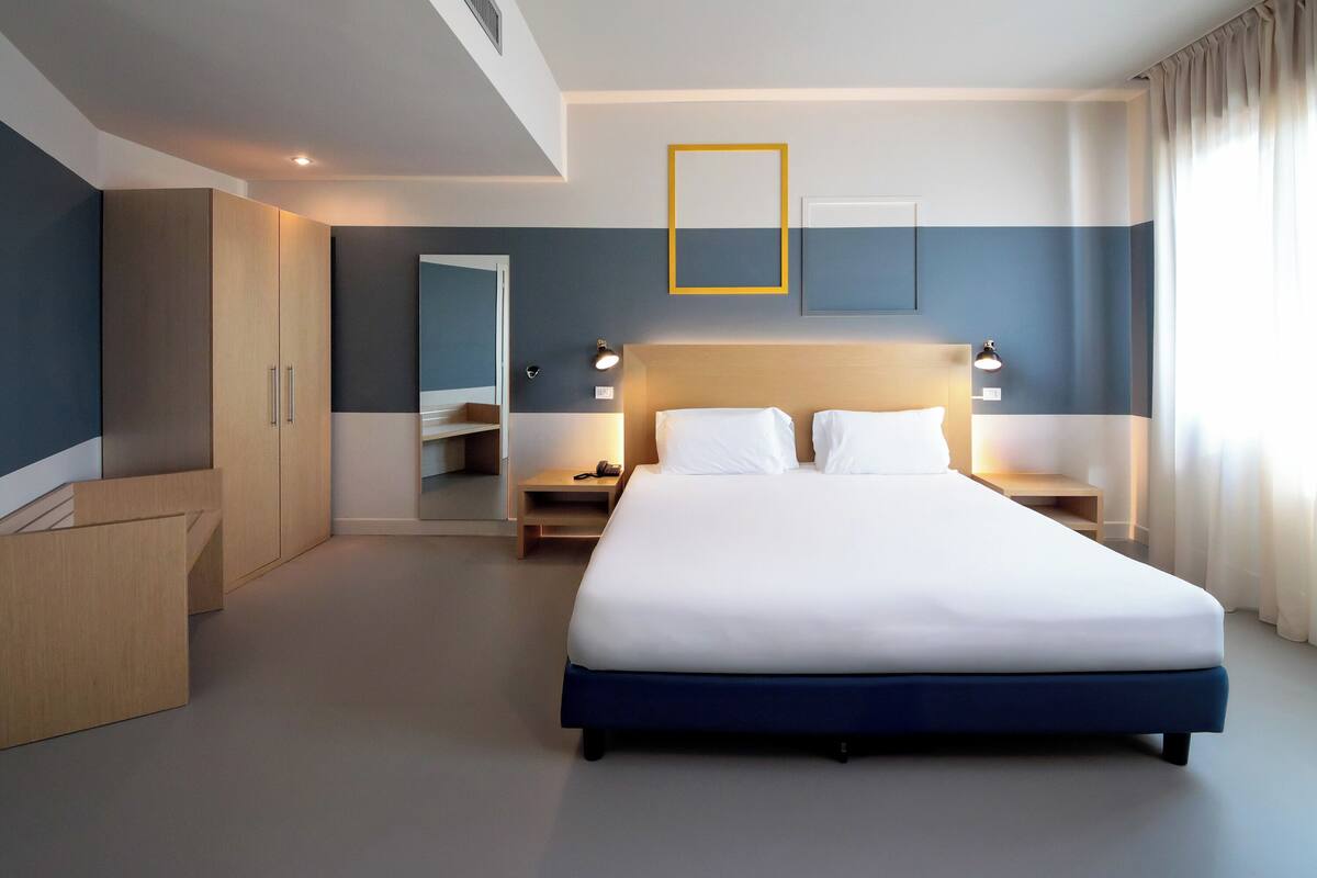 Ein modernes Hotelzimmer mit einem großen Bett, einem Holzschrank, Nachttischen, einem Spiegel und Blick ins Freie.