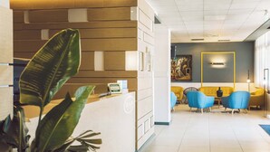 Zona de recepción de un hotel con mostrador, una planta, un cuadro y un área de asientos con sillas.