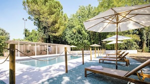 Un área de piscina con sillones de descanso y sombrillas, rodeada de árboles.