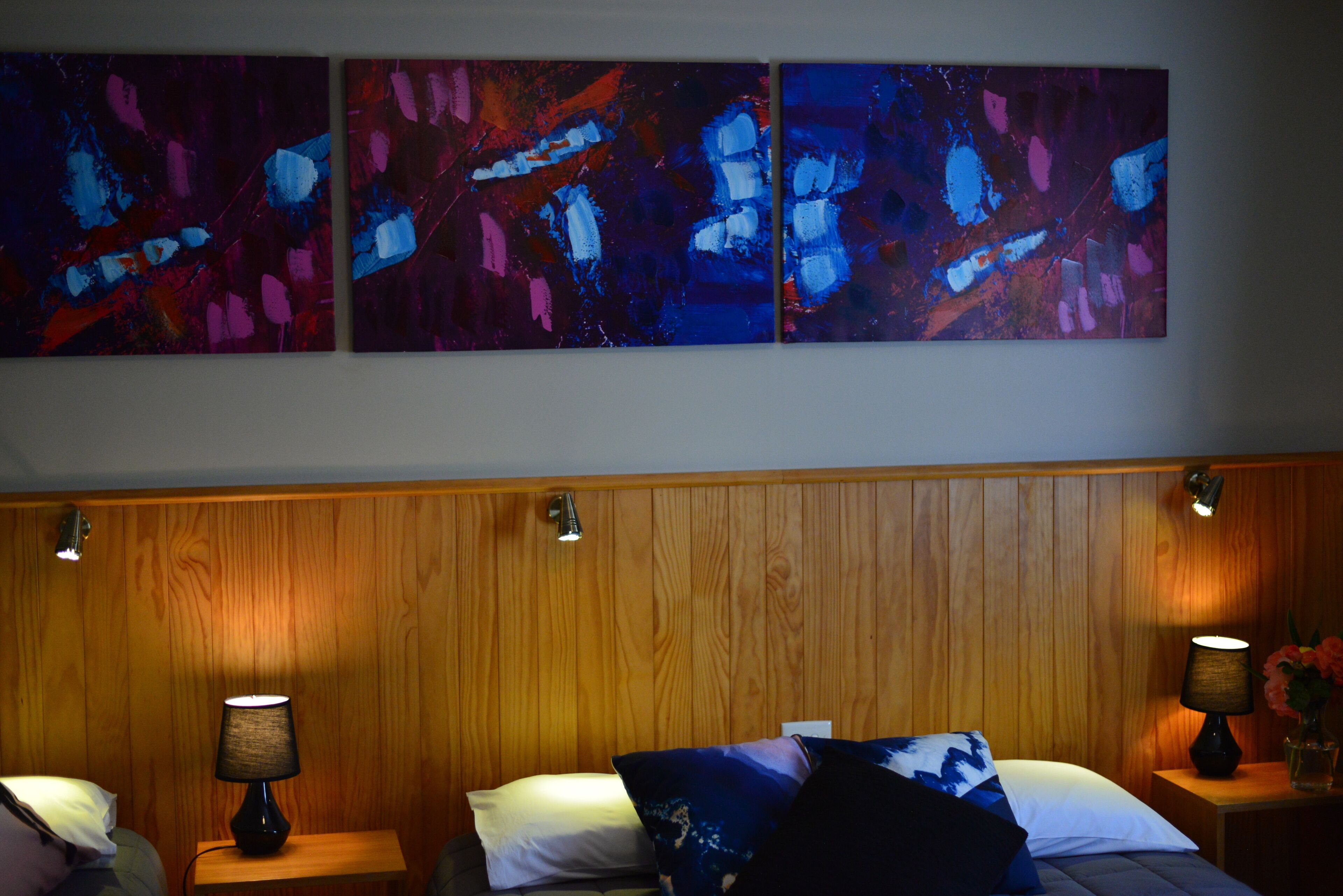 Ein Hotelzimmer mit Holzwandverkleidung, zwei Nachttische mit Lampen, ein Bett mit Kissen und drei abstrakte Gemälde an der Wand.