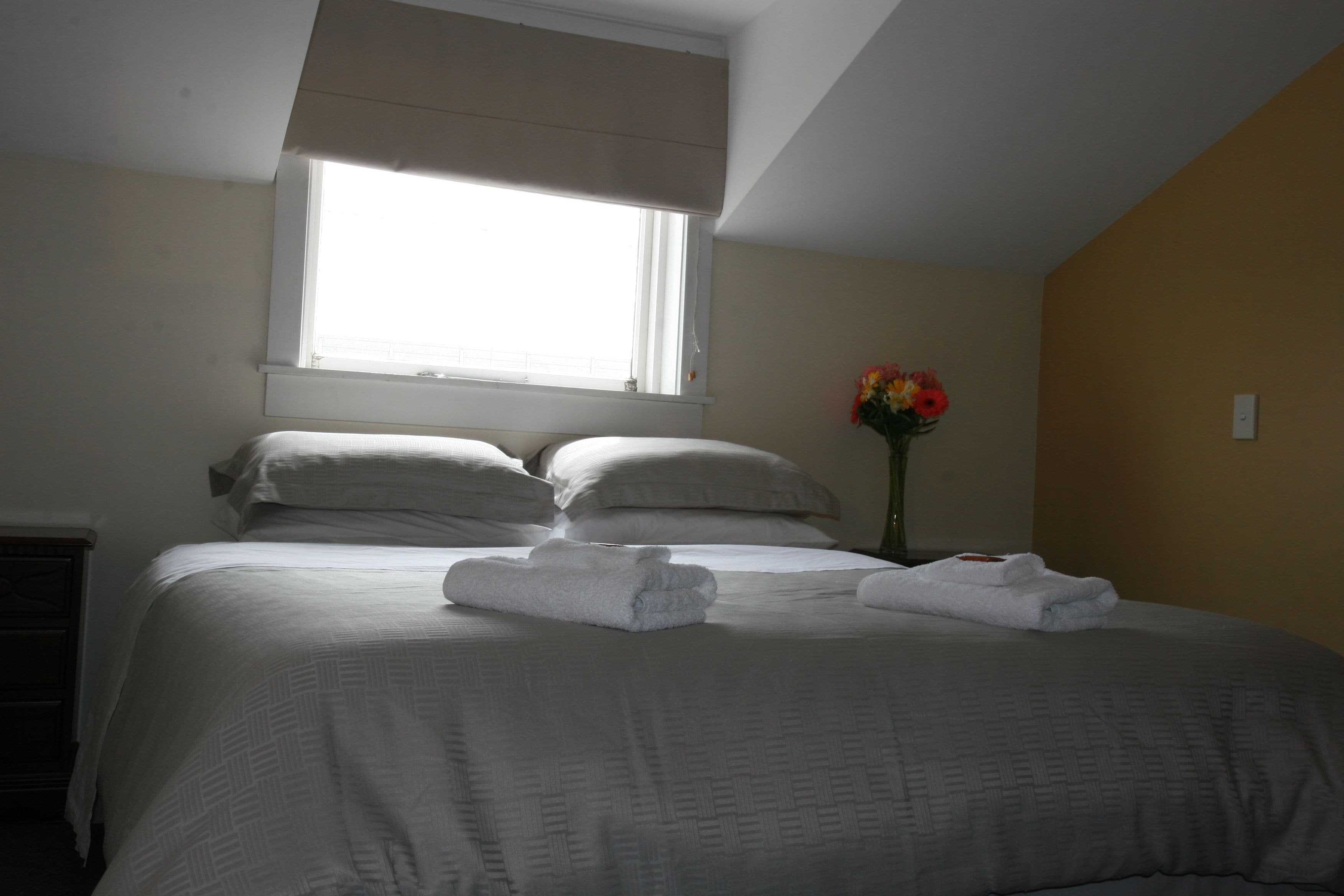 Una cama bien tendida con sábanas y almohadas blancas, un florero con flores en la mesita de noche, y una ventana que deja entrar luz natural.