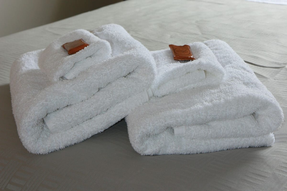Cama bem arrumada com lençóis brancos e um par de toalhas dobradas com etiquetas marrons em um quarto de hotel.
