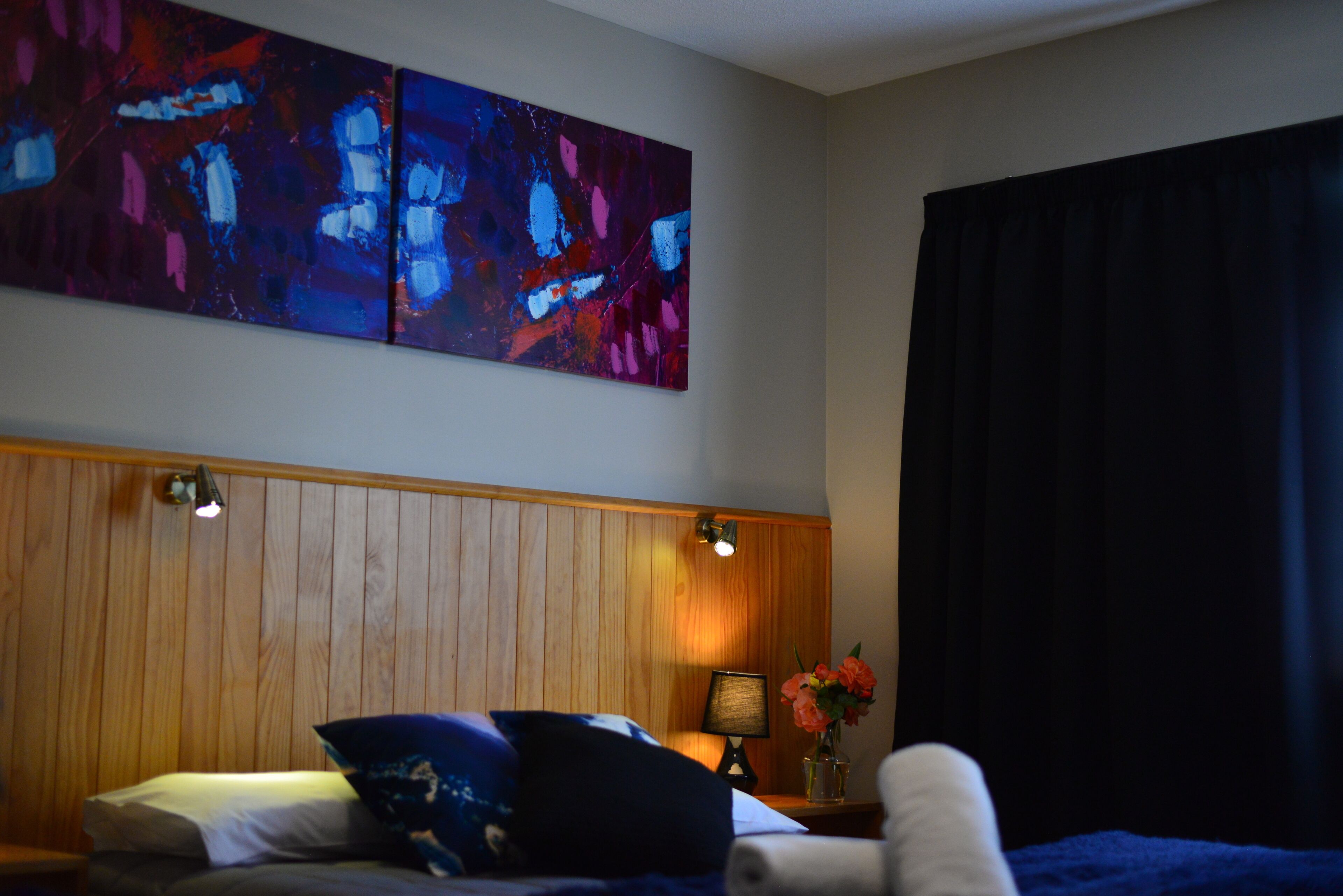 Ein Hotelzimmer mit einem hölzernen Kopfteil, einem Bett mit Kissen, einem Nachttisch mit Lampe und einem Bild an der Wand.