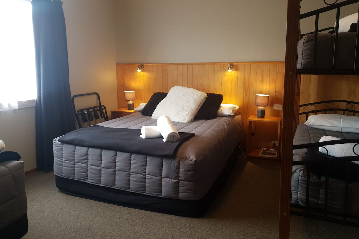 Quarto de hotel com uma cama grande, duas mesas de cabeceira com abajures, um painel de cama de madeira e uma cama beliche no canto.
