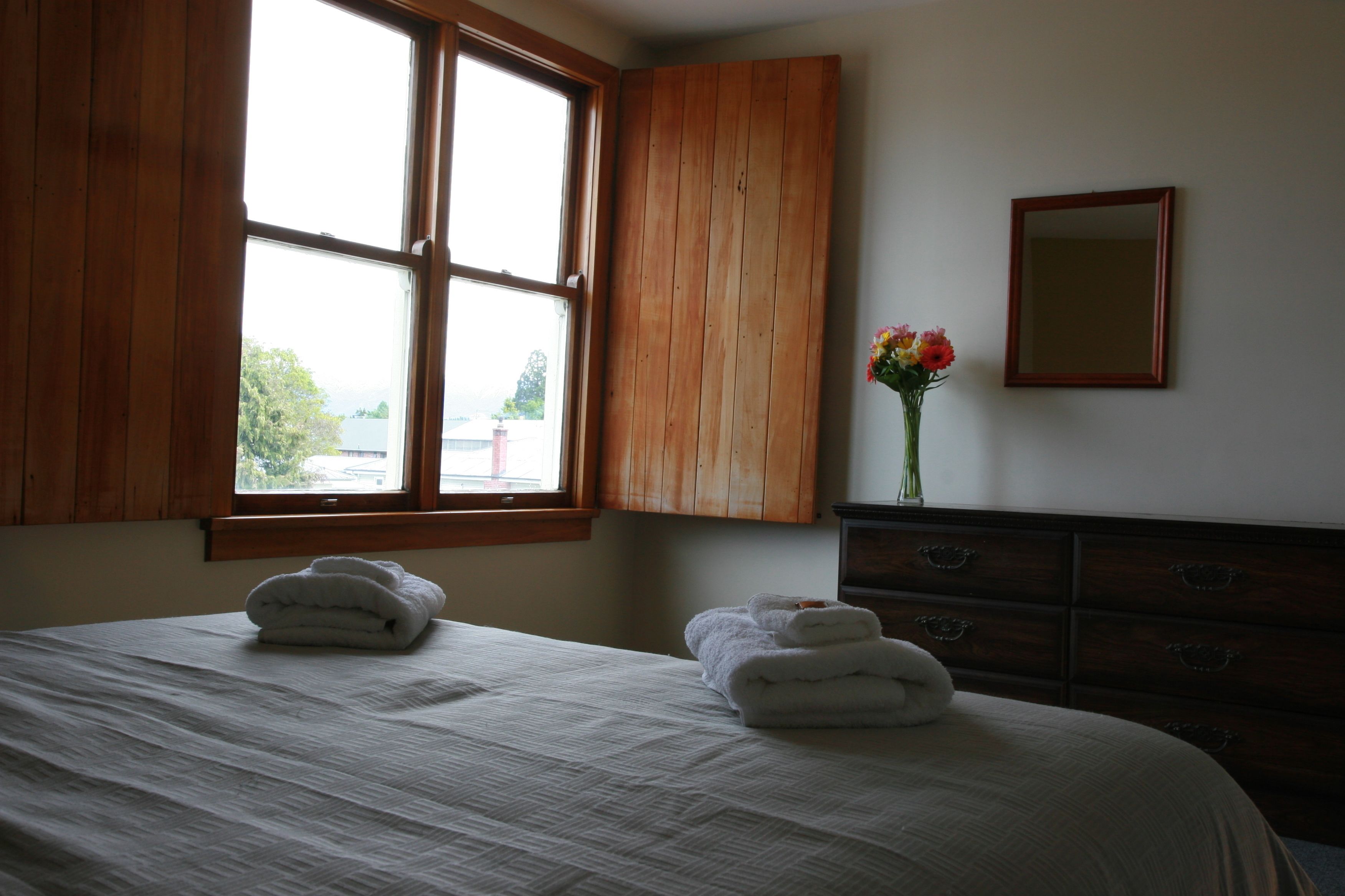 Ein Schlafzimmer mit großem Fenster, Holzschrank, Spiegel und einer Vase mit Blumen.