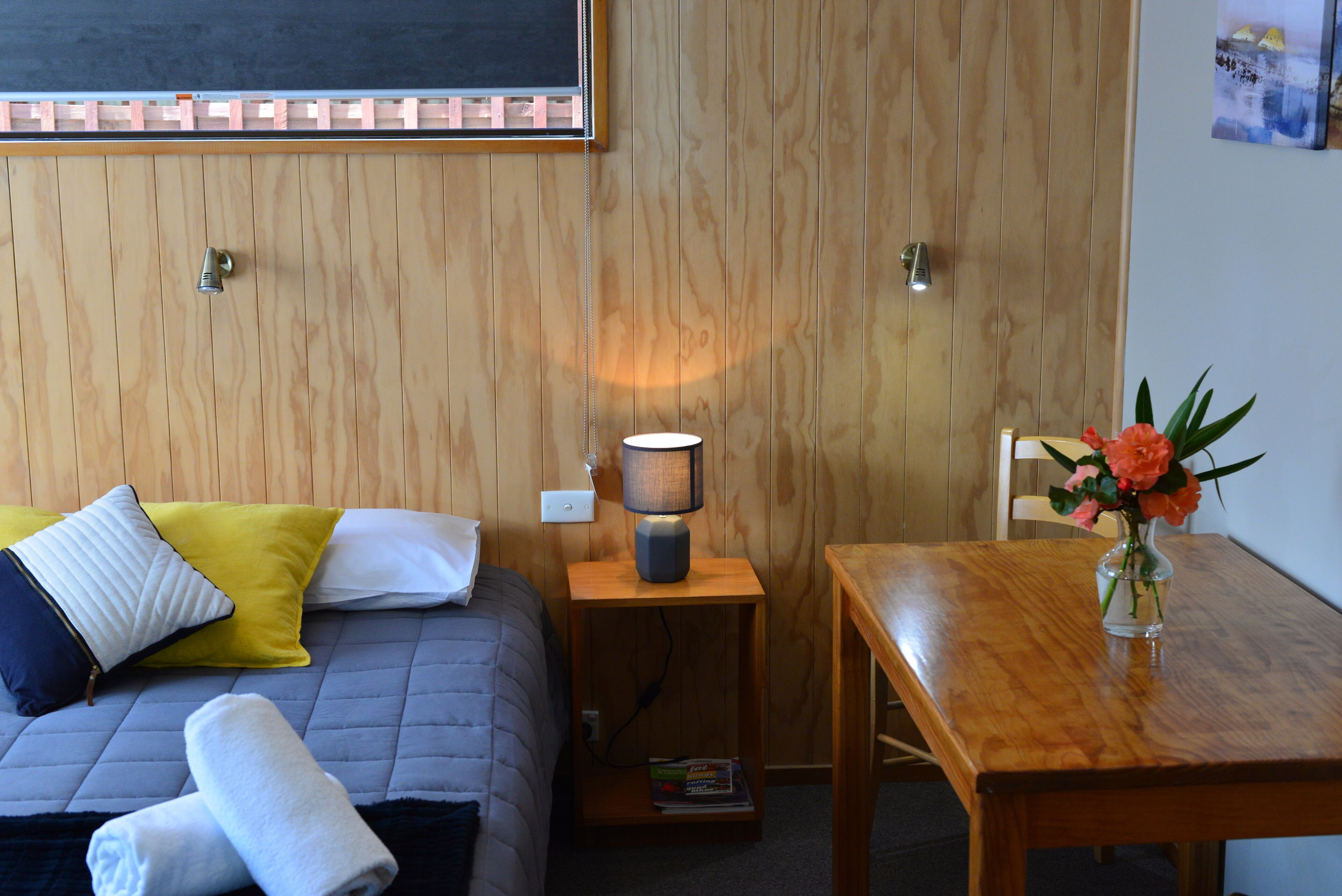 A wooden-paneled room with a bed, bedside table with a lamp, a small table with a vase of flowers, and a chair.