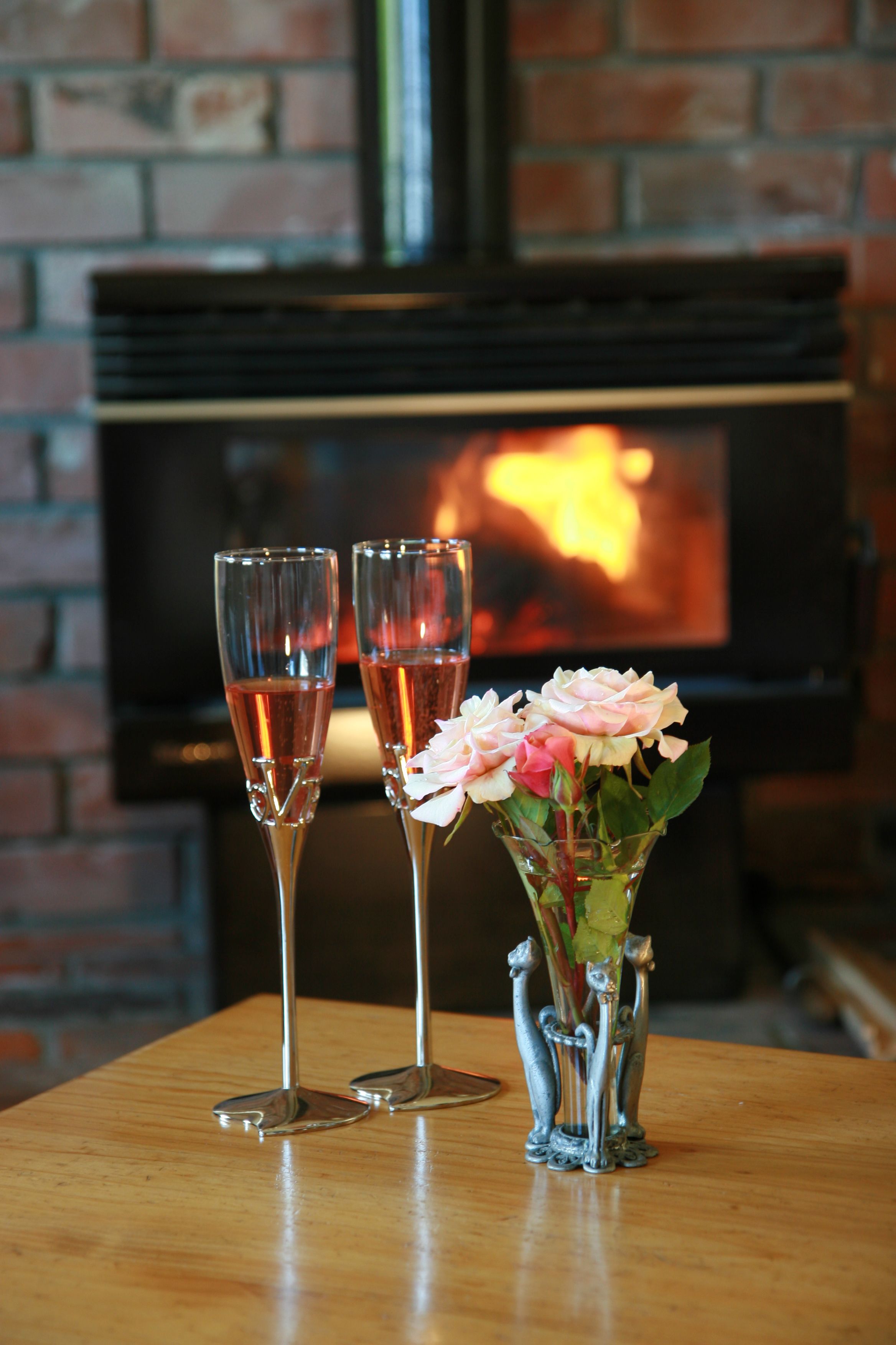Twee champagneglazen en een vaas met bloemen op een houten tafel, met een bakstenen muur en een open haard op de achtergrond.