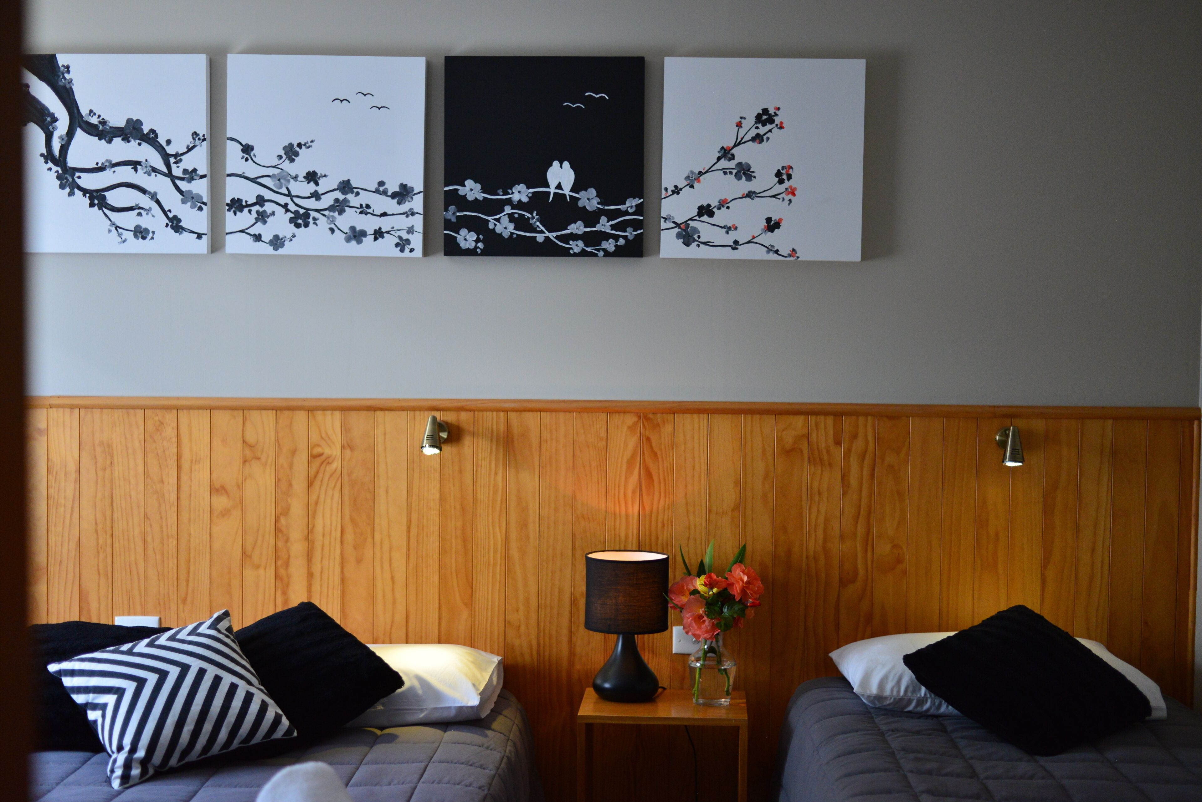 Habitación de hotel con dos camas, una mesita de noche con lámpara y florero, y tres obras de arte murales que representan paisajes naturales.