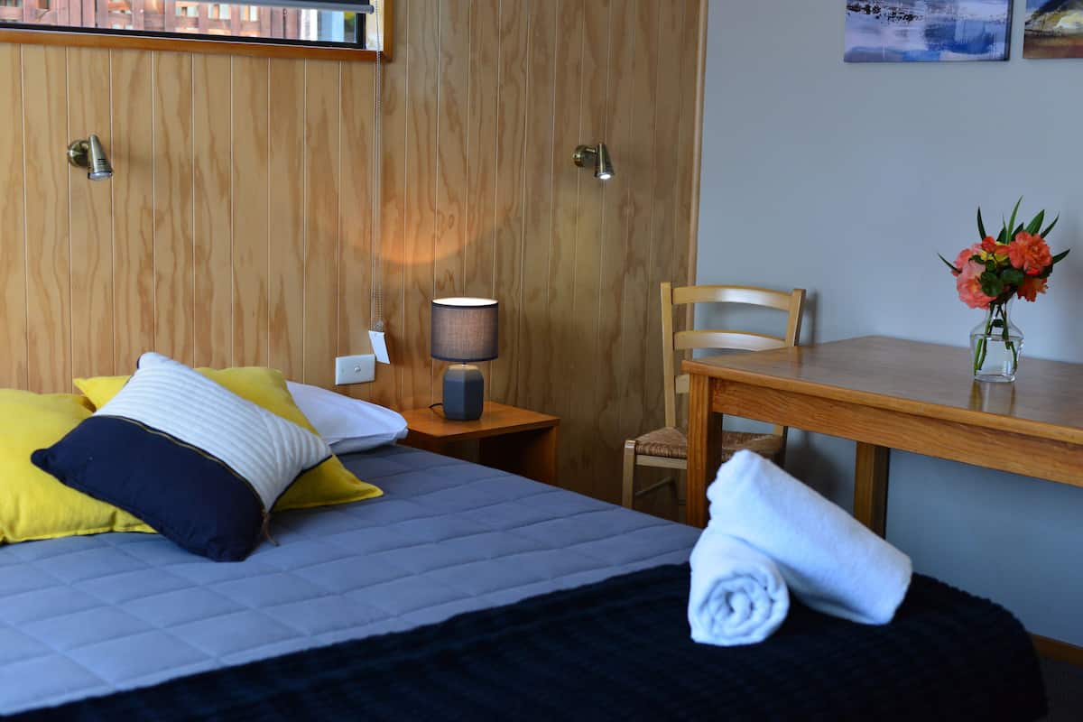 Quarto de hotel com cama, mesa de cabeceira de madeira com abajur, cadeira e mesa com um vaso de flores.