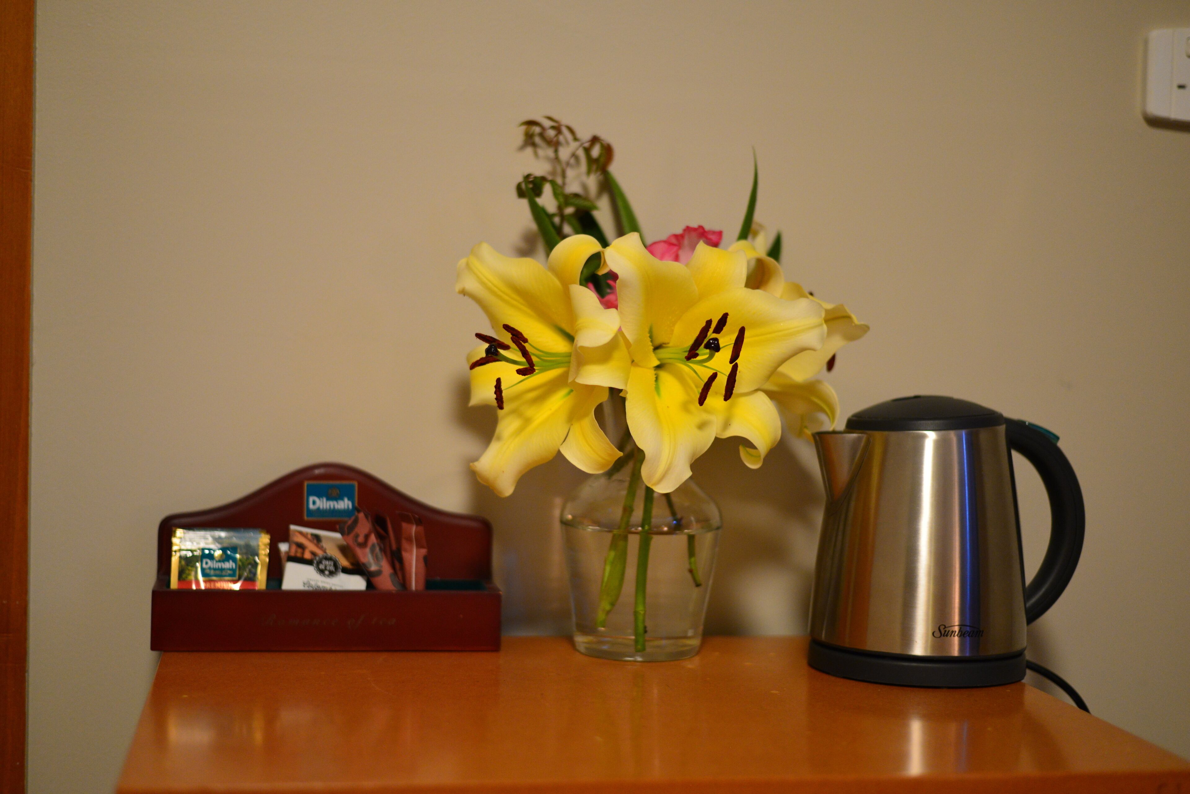 A wooden table with a clear glass vase containing yellow flowers, a metal kettle, and a red box with a brand name.
