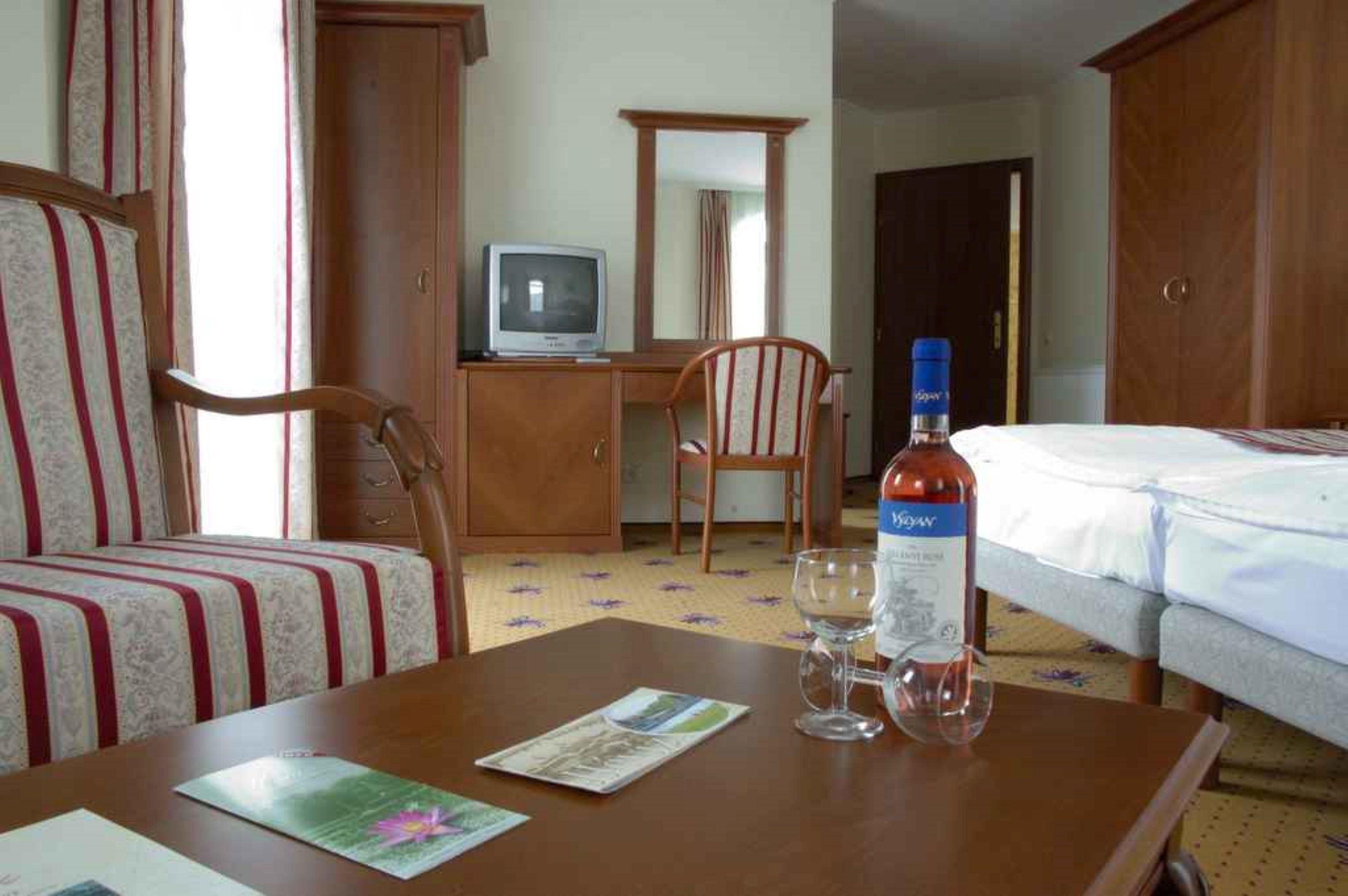 A hotel room with a bed, a desk with a television, a chair, and a bottle of wine.