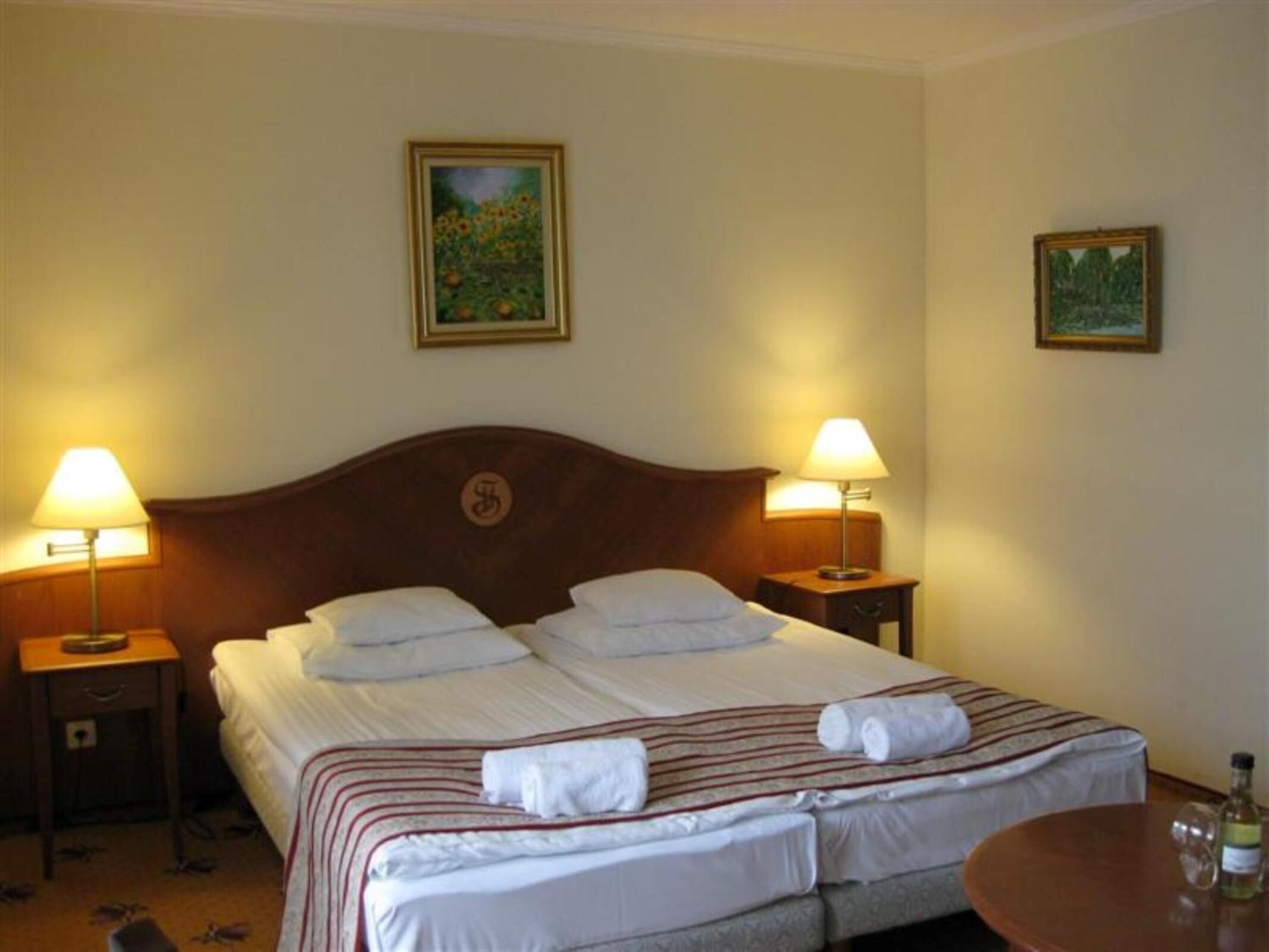A double bed with two pillows, a wooden headboard, two bedside lamps, a small table with a bottle, and two framed pictures on the wall.