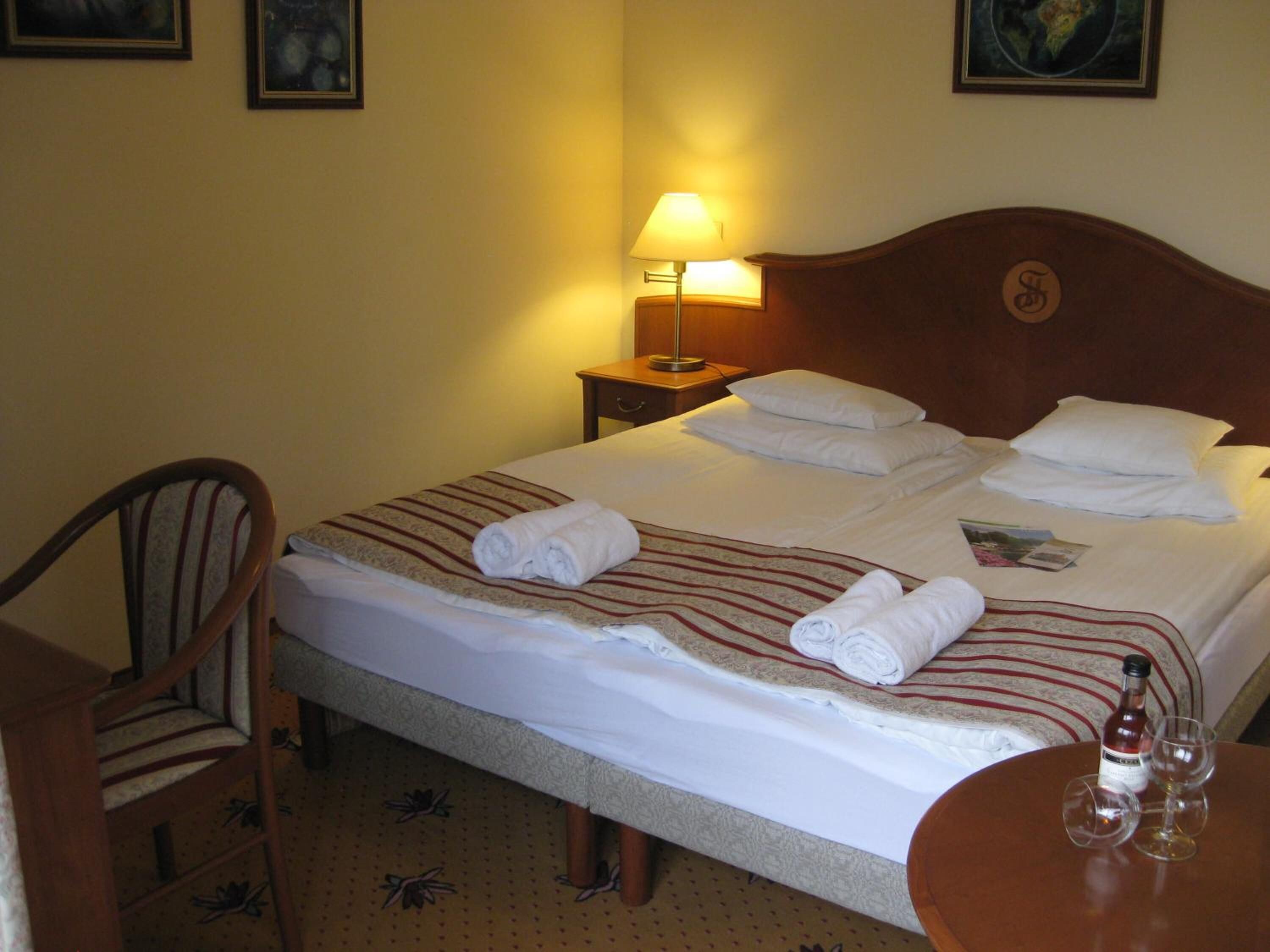 A hotel room with a neatly made bed, a chair, a nightstand with a lamp, and a table with glasses and a bottle.