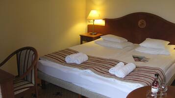 A hotel room with a neatly made bed, a chair, a nightstand with a lamp, and a table with glasses and a bottle.
