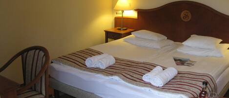 A hotel room with a neatly made bed, a chair, a nightstand with a lamp, and a table with glasses and a bottle.