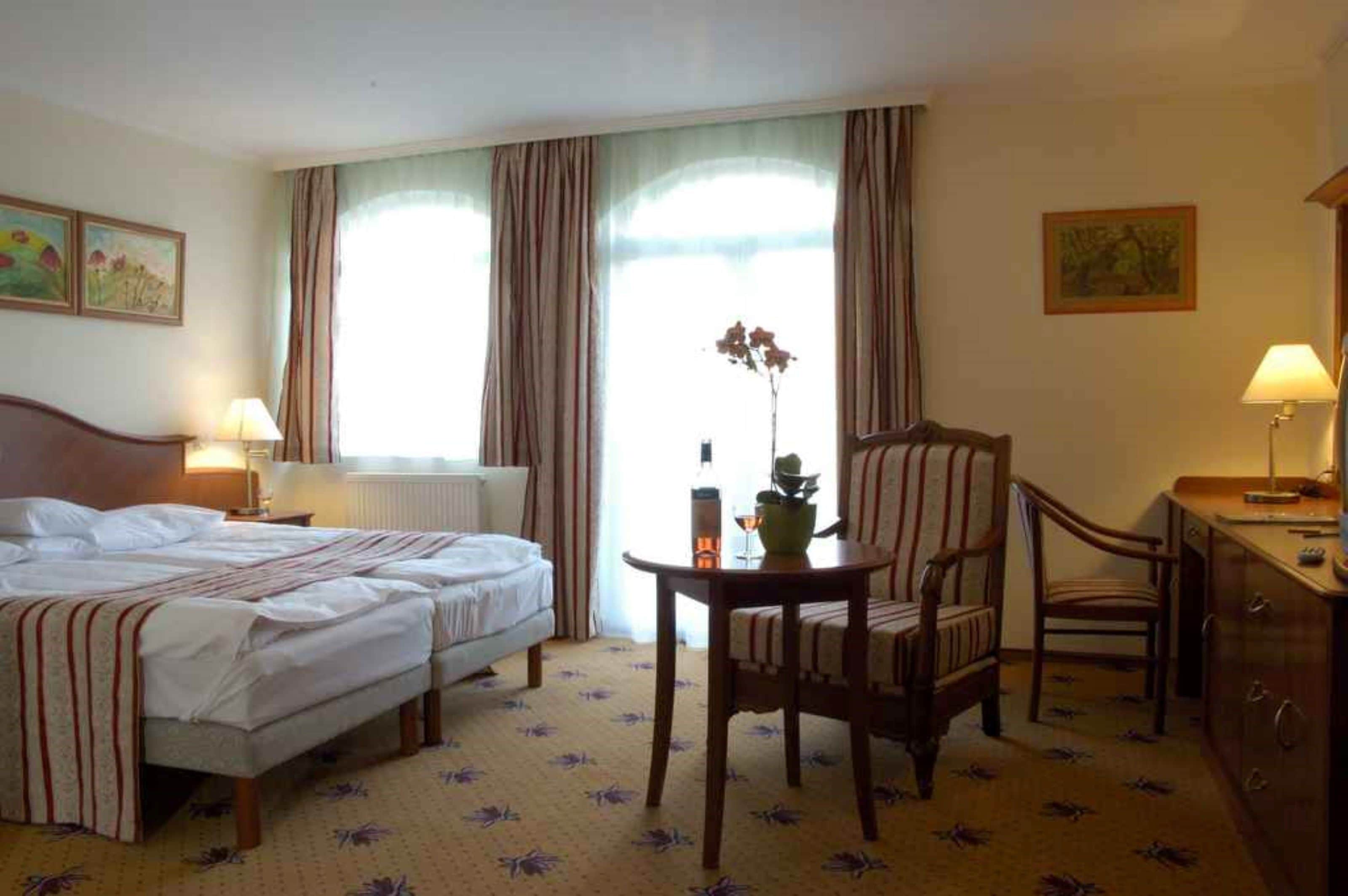 A hotel room with two beds, a small table with a vase, a chair, and a desk with a lamp.