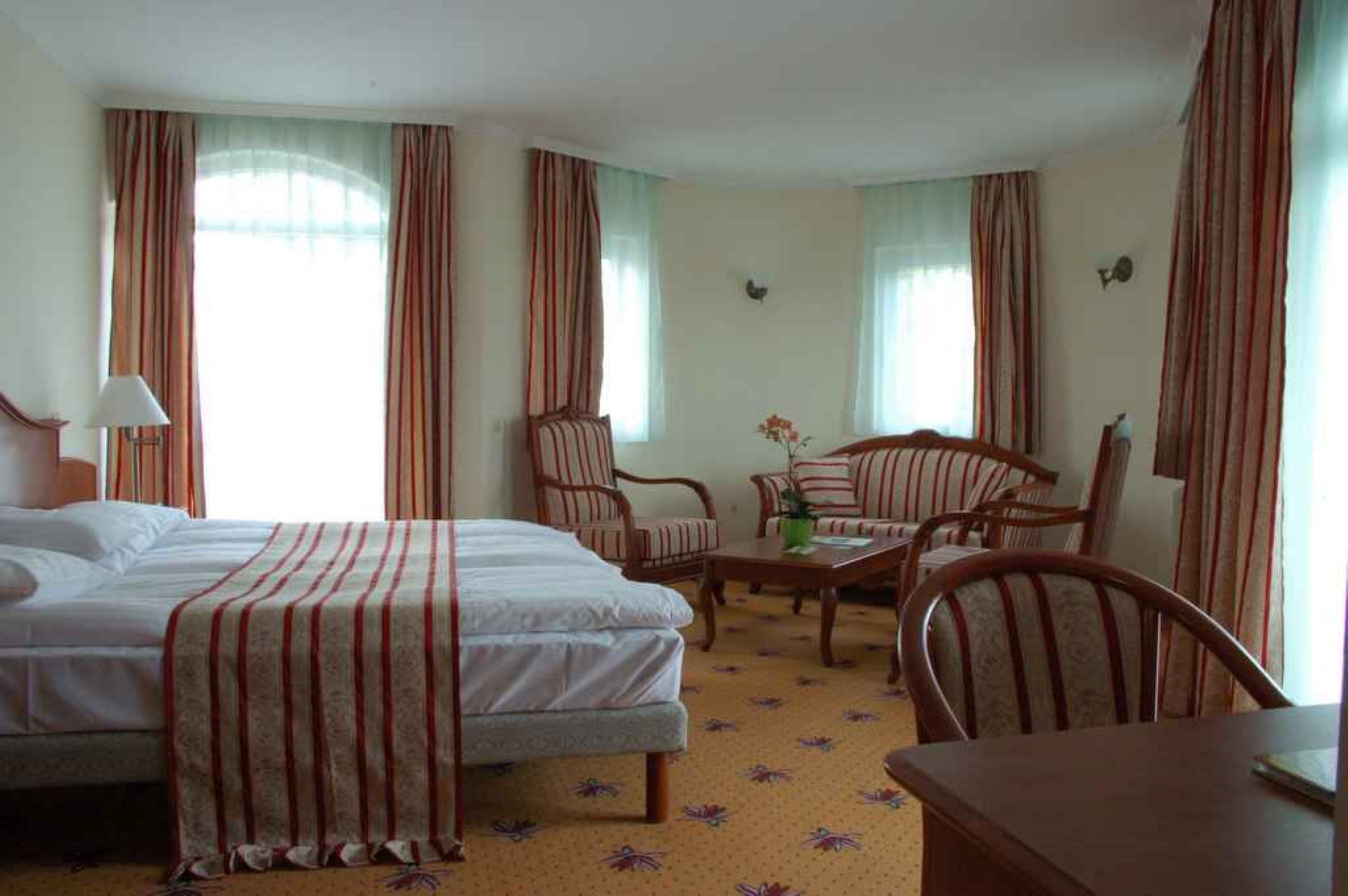 Une chambre d’hôtel avec deux lits, un coin salon avec une table et des chaises, et de grandes fenêtres avec des rideaux.